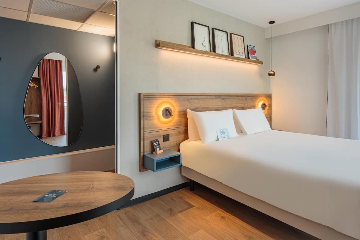 Une chambre d’hôtel avec un grand lit, une table ronde, un miroir et une étagère murale ornée de tableaux encadrés.