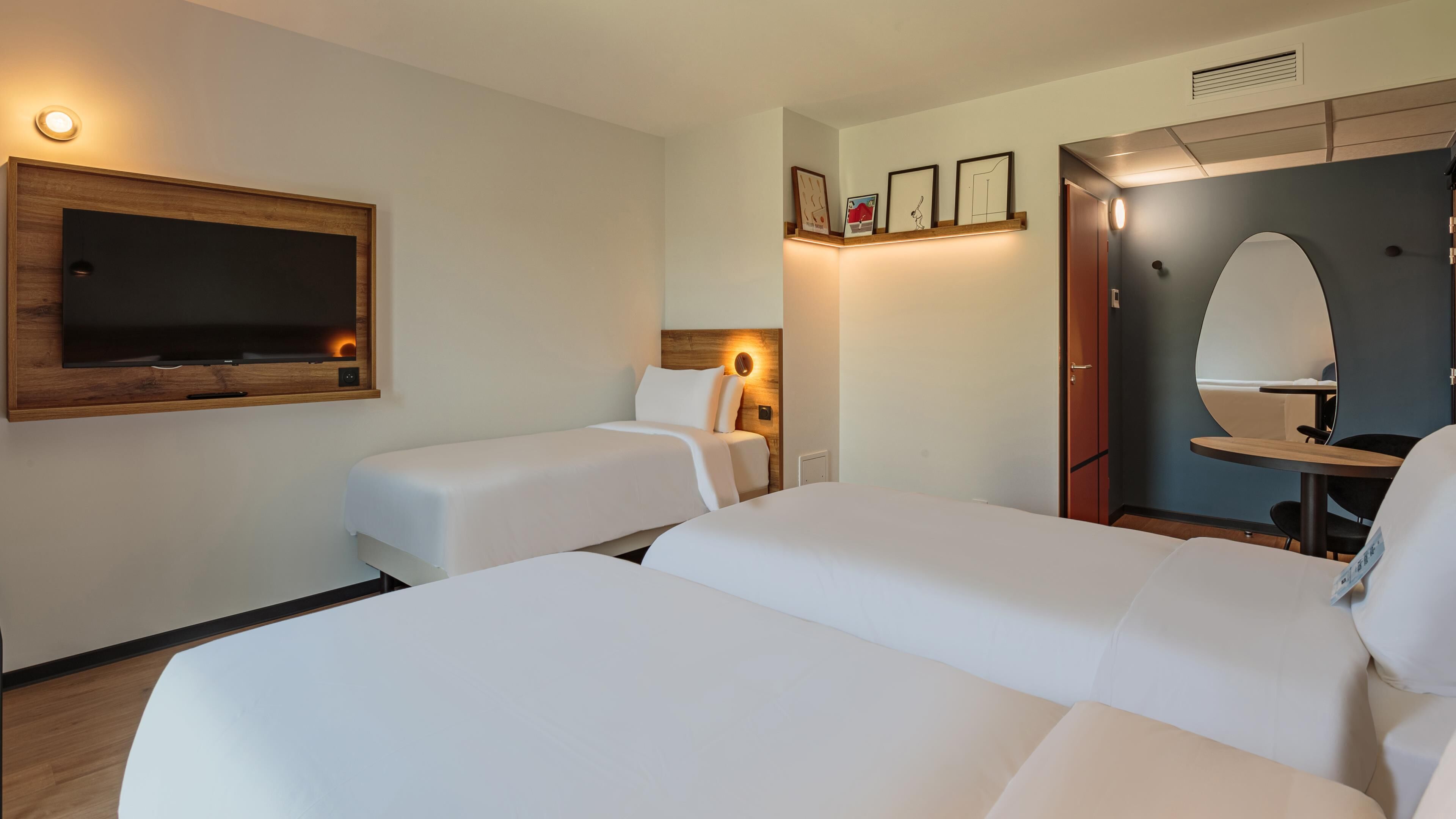 Habitació d'hotel amb dos llits individuals, un televisor de pantalla plana fixat a un panell de fusta, un taulell petit amb una cadira i un mirall a la paret.