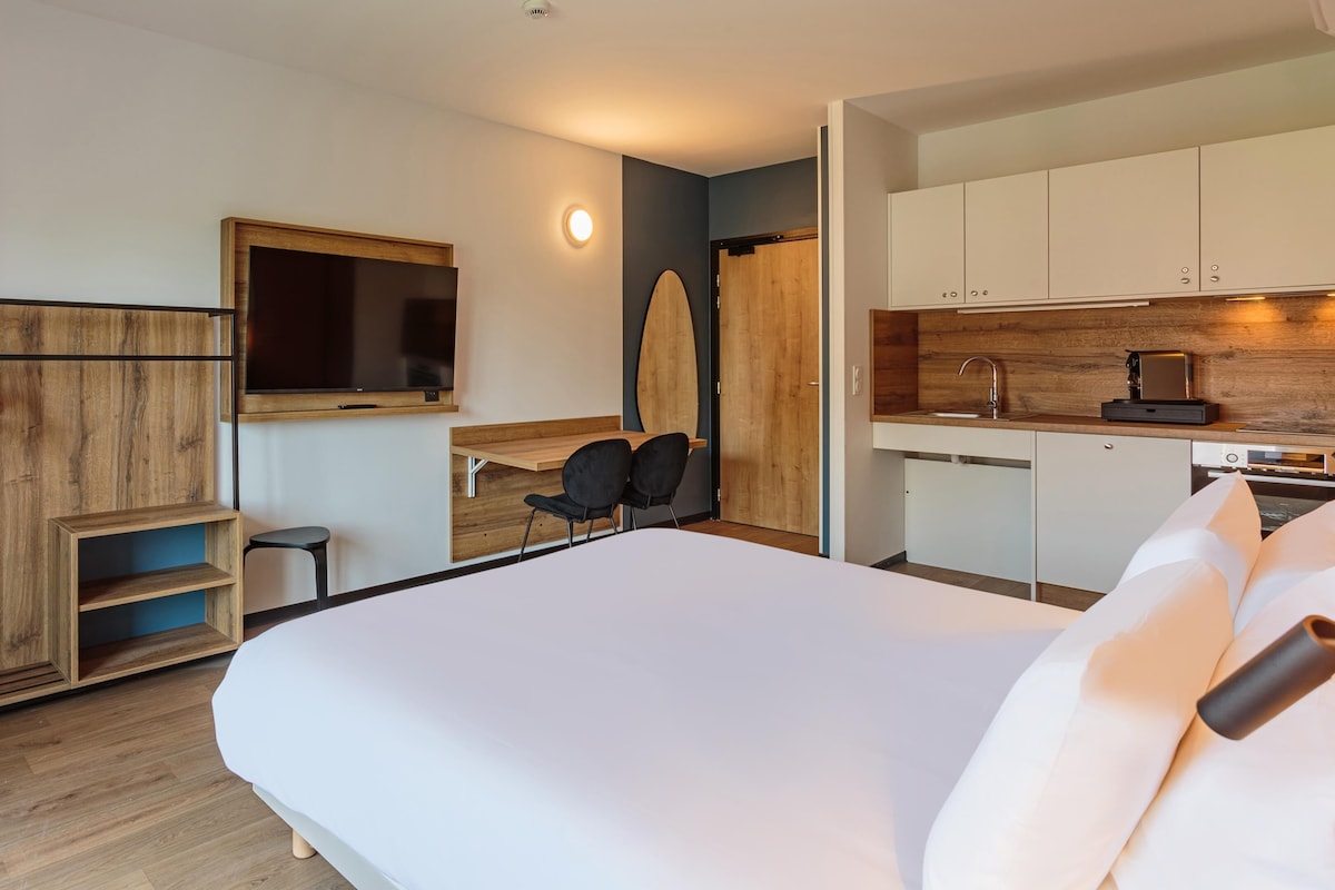 A modern hotel room with a large bed, a TV mounted on the wall, a small kitchen area, and a wooden wardrobe.