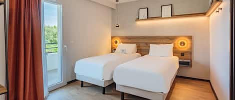 A hotel room with two beds, a sliding glass door to a balcony, and a wooden headboard with wall-mounted lights.