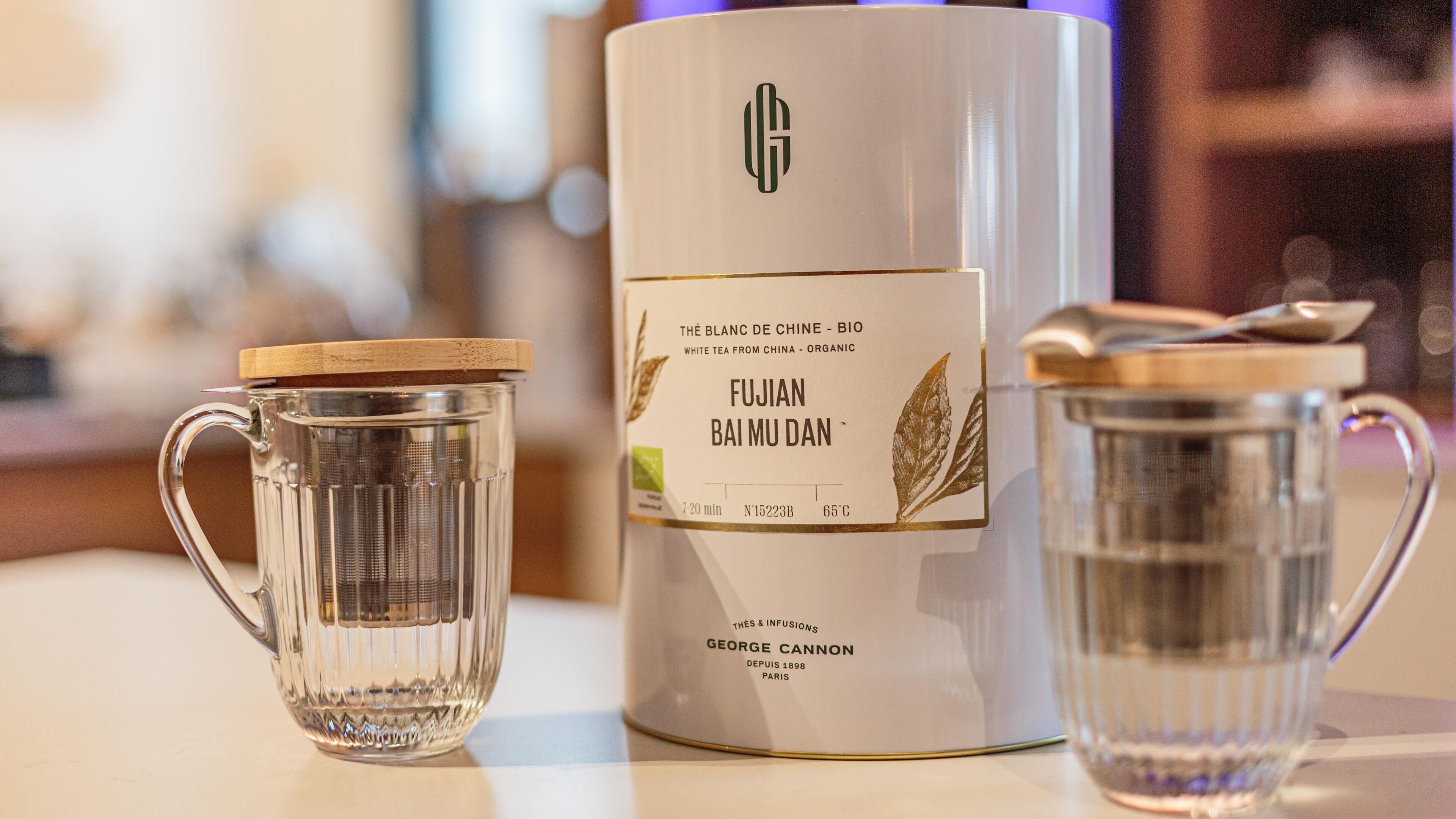 Un juego de té que incluye una tetera y dos tazas de vidrio, con una etiqueta que indica 'FUJIAN BAI MU DAN' y 'THÉ BLANC DE CHINE - BIO'.