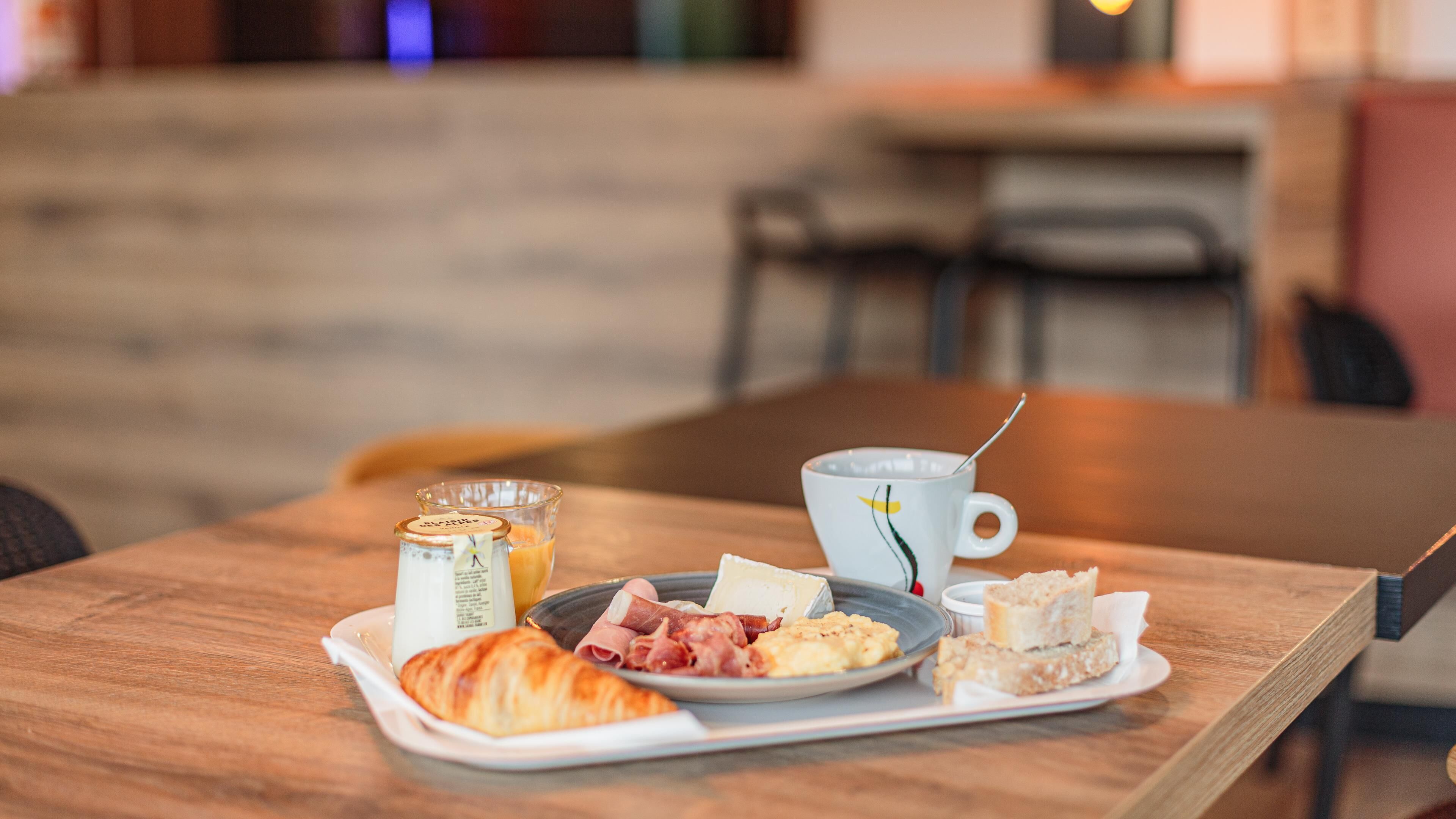 Un plateau de petit-déjeuner avec un croissant, du jambon, des œufs brouillés et une tasse de café, posé sur une table en bois.