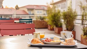A breakfast tray with croissants, ham, scrambled eggs, and toast on a table with a view of a balcony, potted plants, and red chairs.