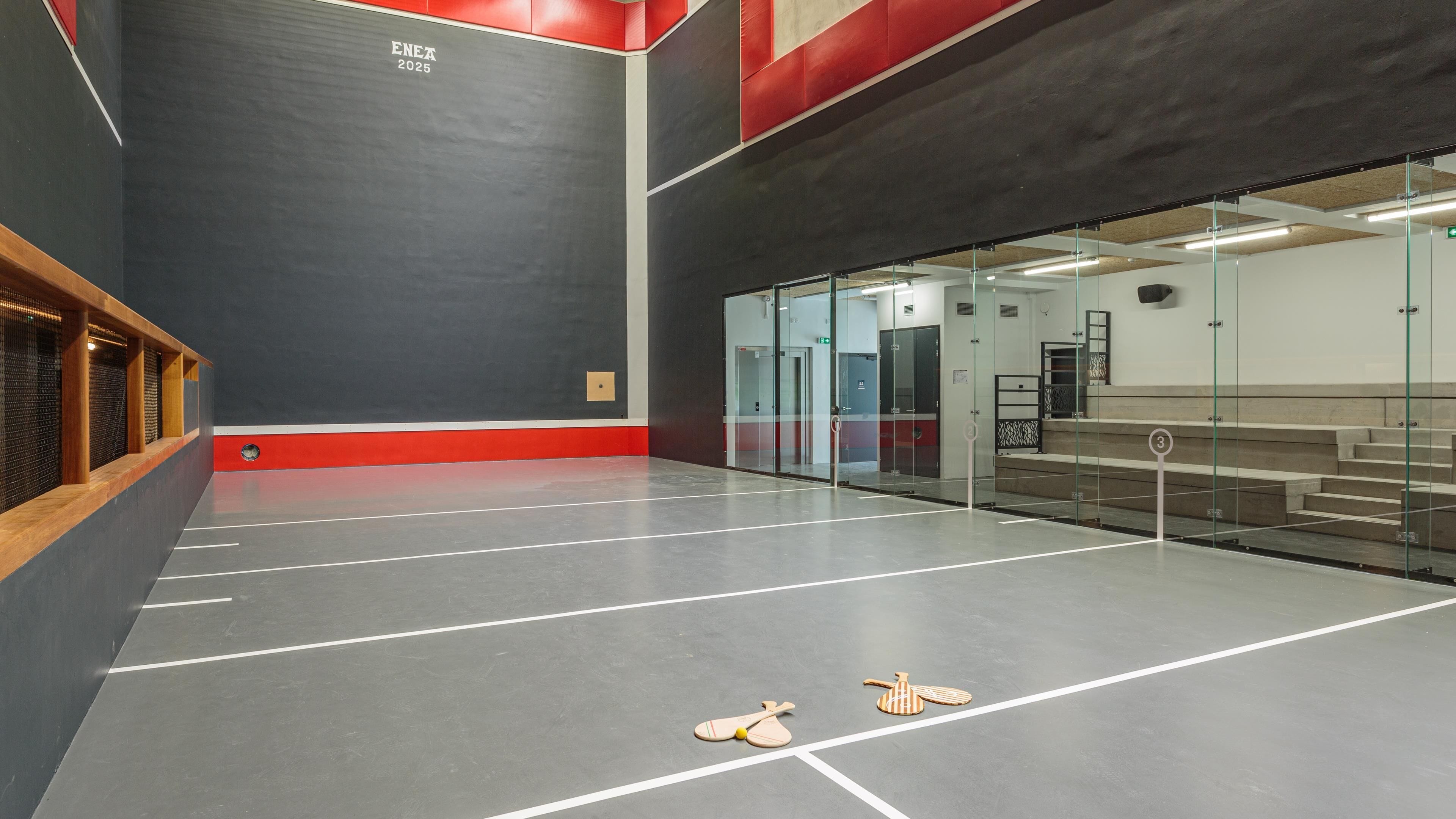 A modern indoor tennis court with a red and black color scheme, glass walls, and a wooden bench area.