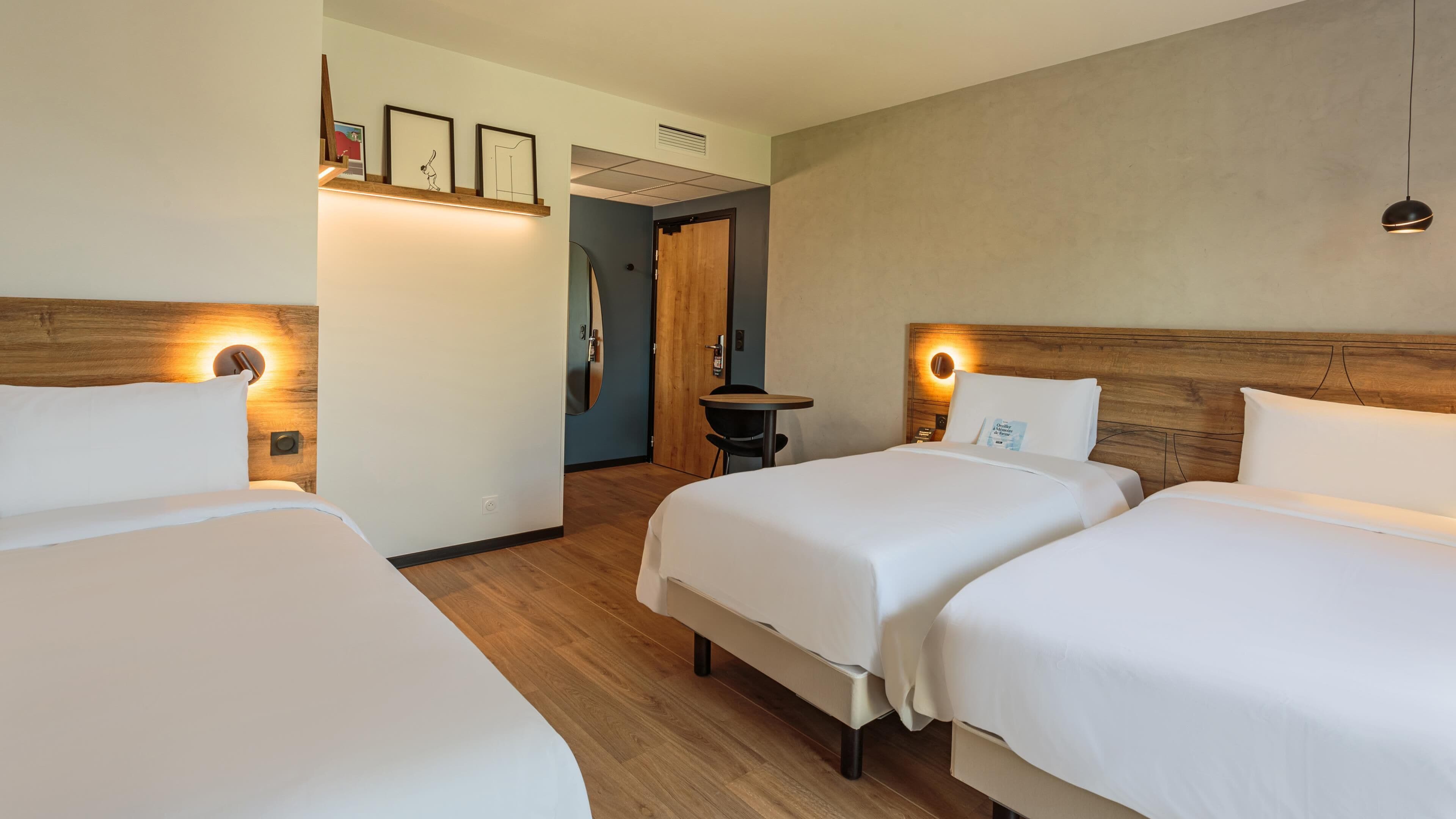 A hotel room with two single beds, a wooden headboard, a small table, and a wall-mounted shelf with framed pictures.