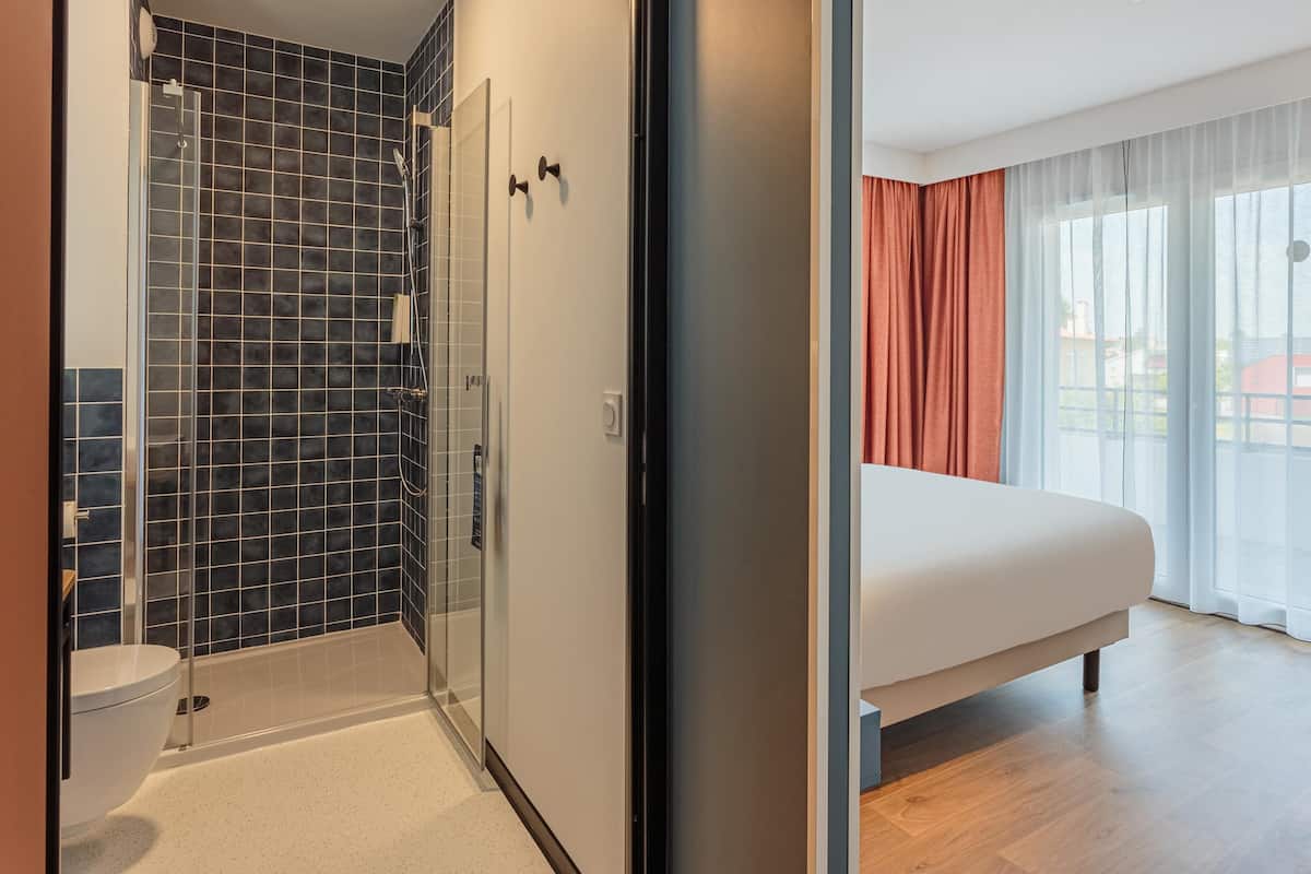 Une chambre d’hôtel dotée d’une douche en verre, d’un lit, d’une grande fenêtre avec des rideaux et offrant une vue sur l’extérieur.