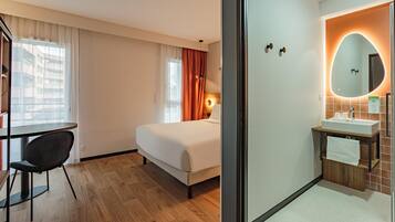 A hotel room with a bed, a desk, a chair, a bathroom with a sink and mirror, and a view of buildings through the window.