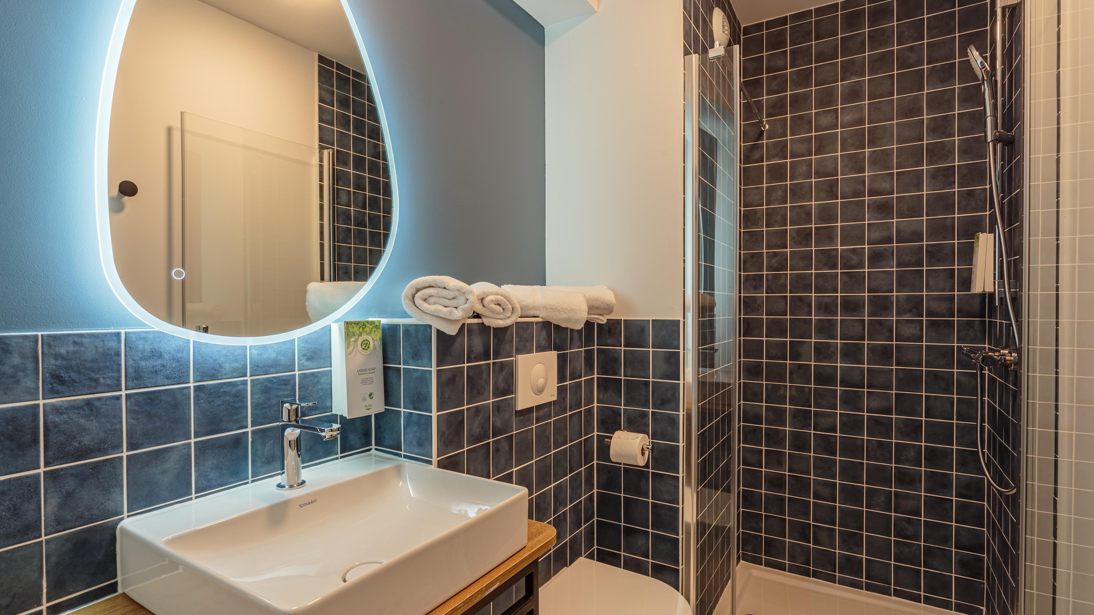 A bathroom with a white sink, a mirror, a towel rack with towels, a shower area with a glass door, and dark tiled walls.
