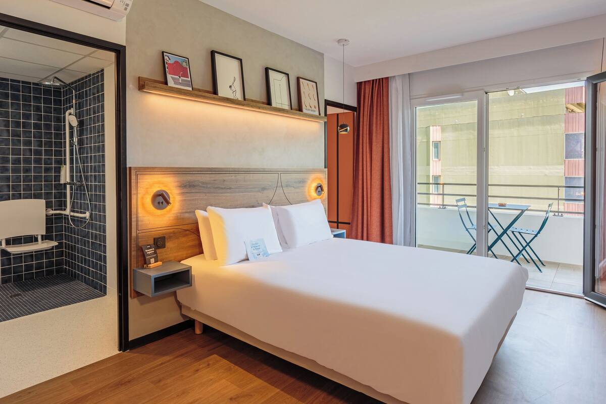 Une chambre d’hôtel avec un grand lit, un espace douche, une table de chevet avec une lampe et une vue sur le balcon extérieur.