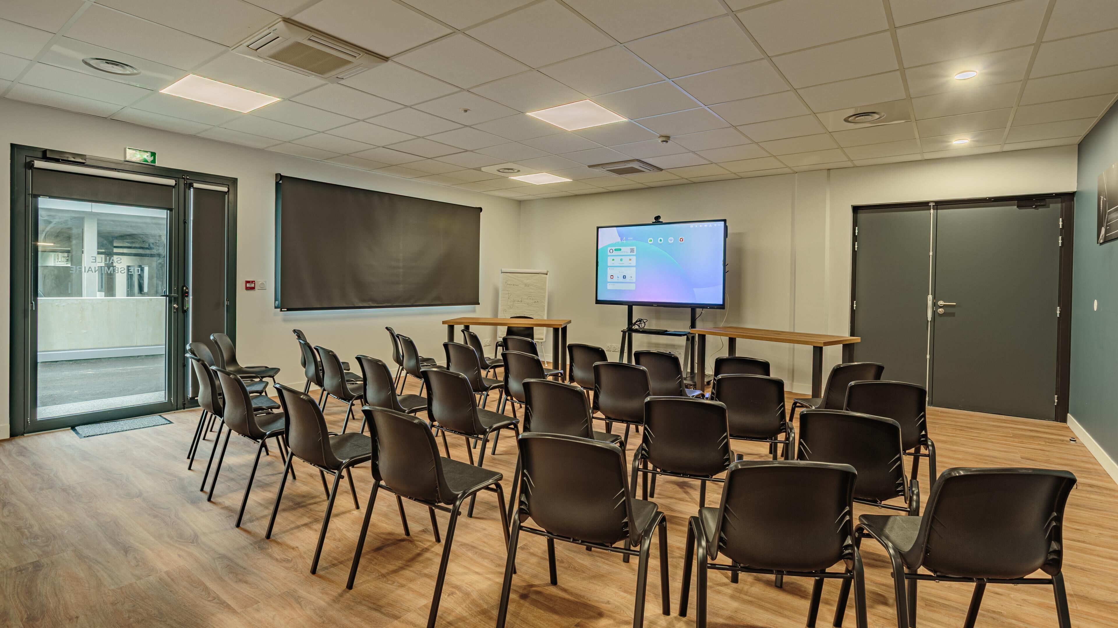 A conference room with a large screen, multiple chairs, a wooden table, and a door with a view of a parking garage.