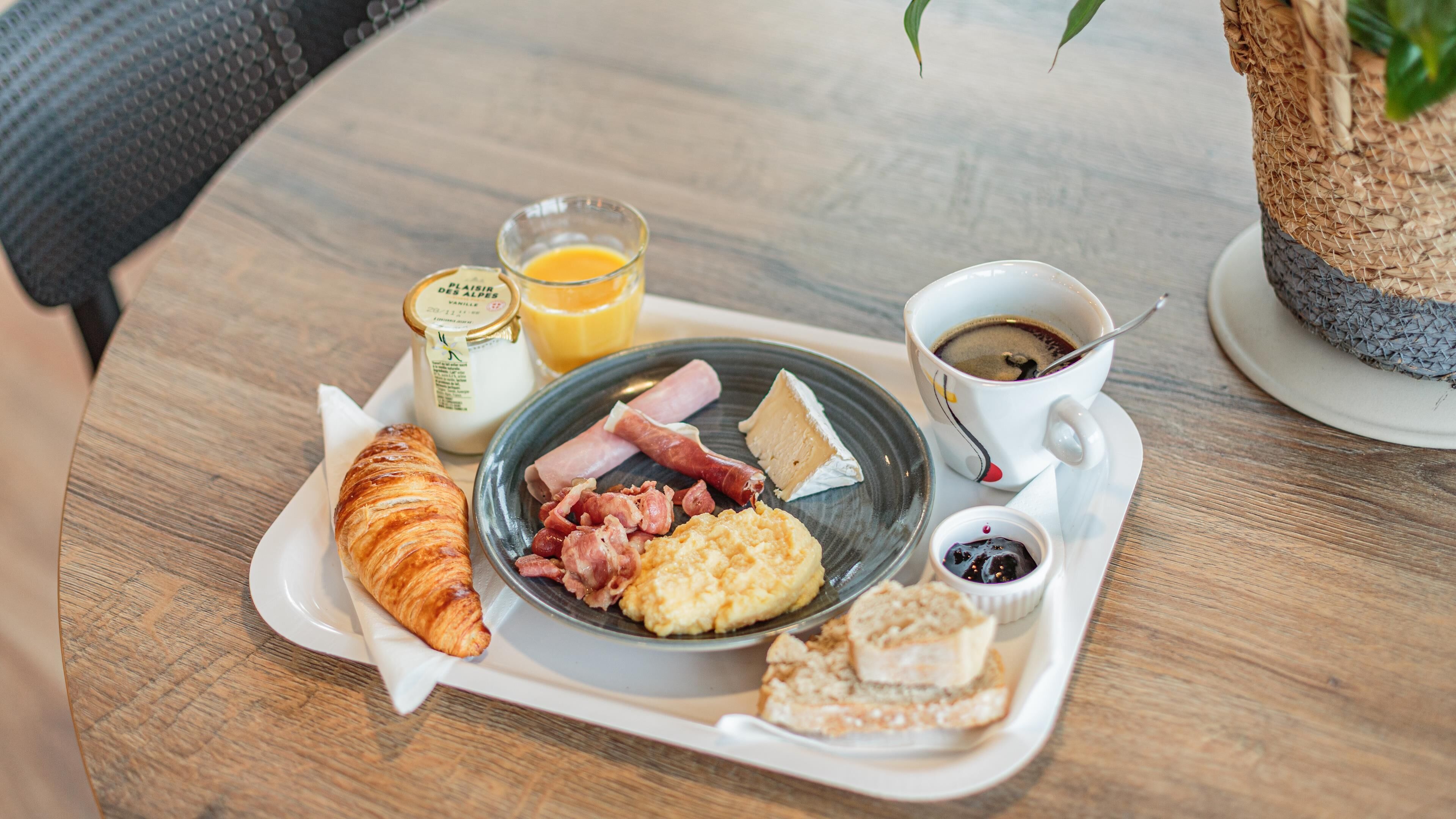 Bandeja de desayuno con croissant, jamón, queso, huevos revueltos, café y jugo de naranja sobre una mesa de madera.
