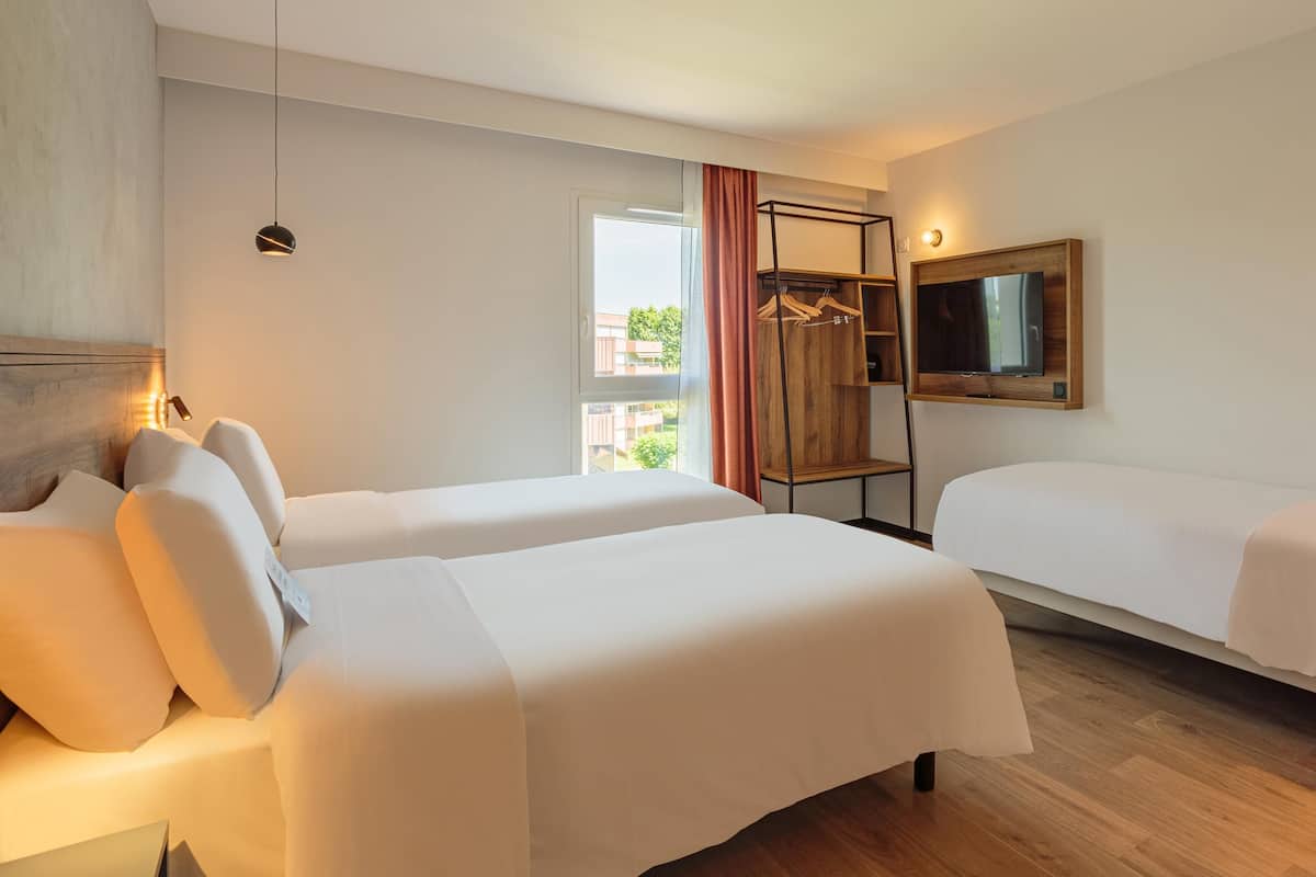 Une chambre d’hôtel avec deux lits, un téléviseur fixé au mur, un plancher en bois et une fenêtre avec des rideaux.