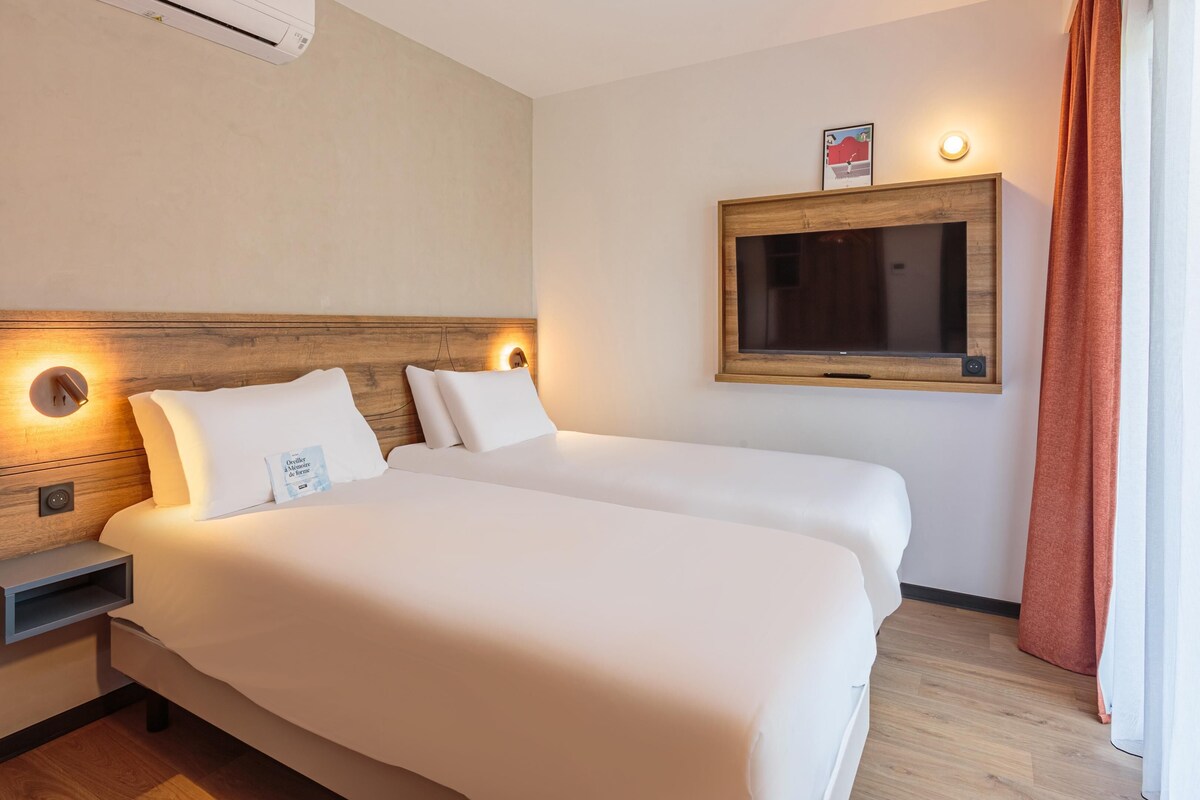 A hotel room with two beds, a wooden headboard, a bedside table, a TV mounted on the wall, and a window with curtains.