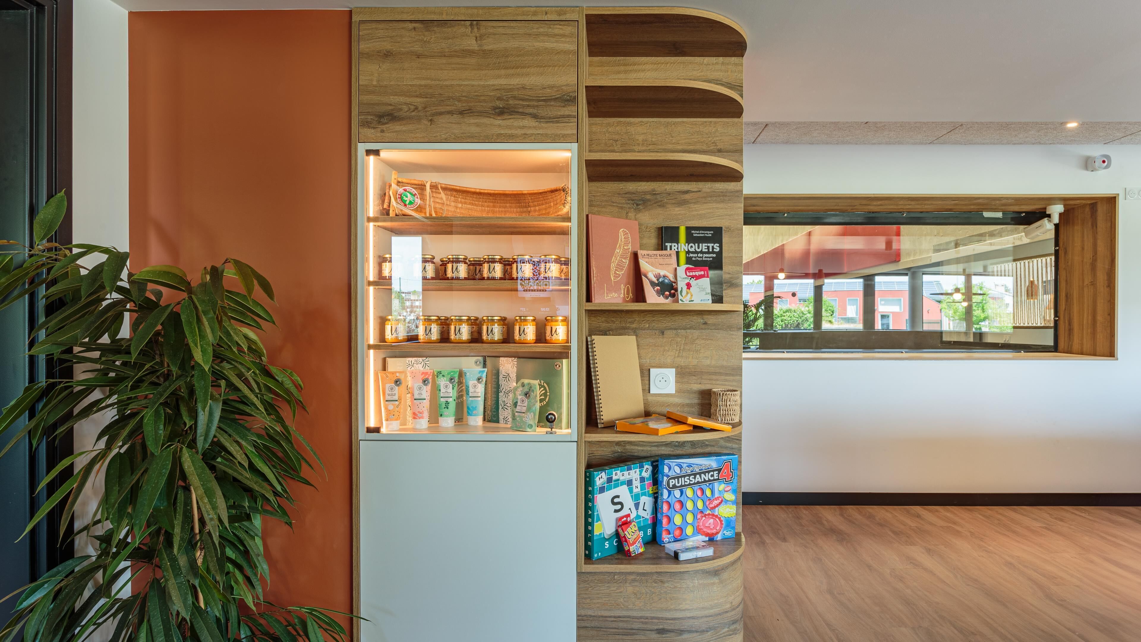 A modern interior with a wooden display unit containing various products, a potted plant, and a view of a red building outside.