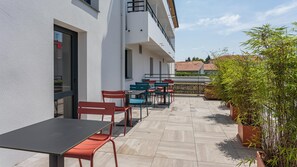 Une terrasse extérieure avec des tables et des chaises, un pot de fleurs et une vue sur un immeuble doté d’un balcon.
