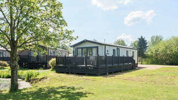 A mobile home with a fenced deck, surrounded by grass and trees.