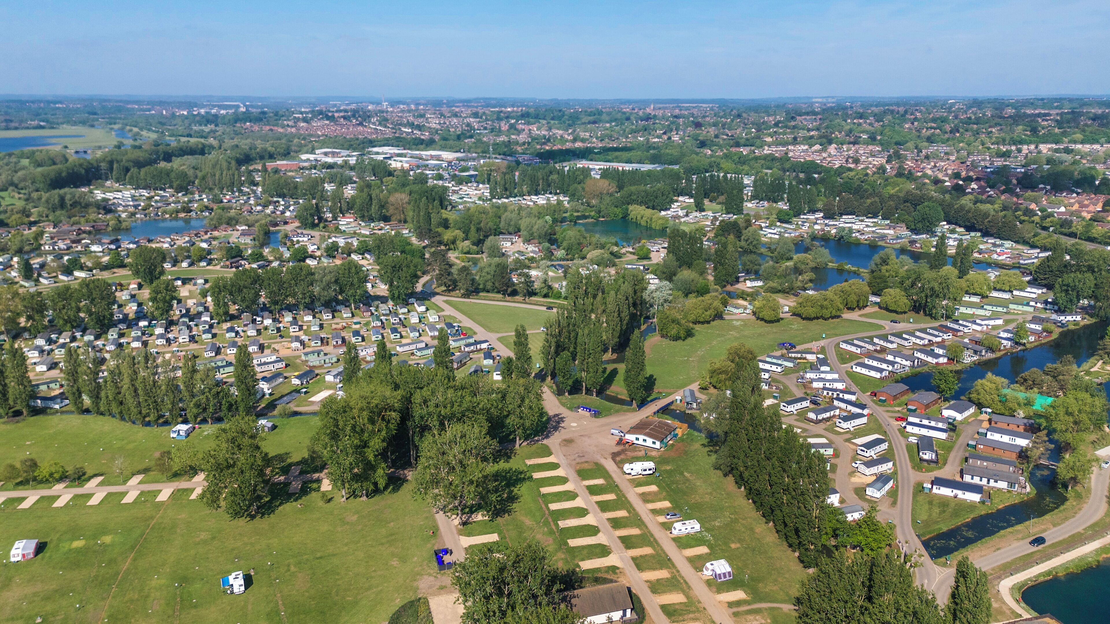 Vista aérea de un camping con vehículos recreativos y casas móviles, rodeado de campos verdes y lagos.