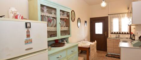 A kitchen with a refrigerator, a cabinet with glass doors, a dining table with a chair, and a window with curtains.