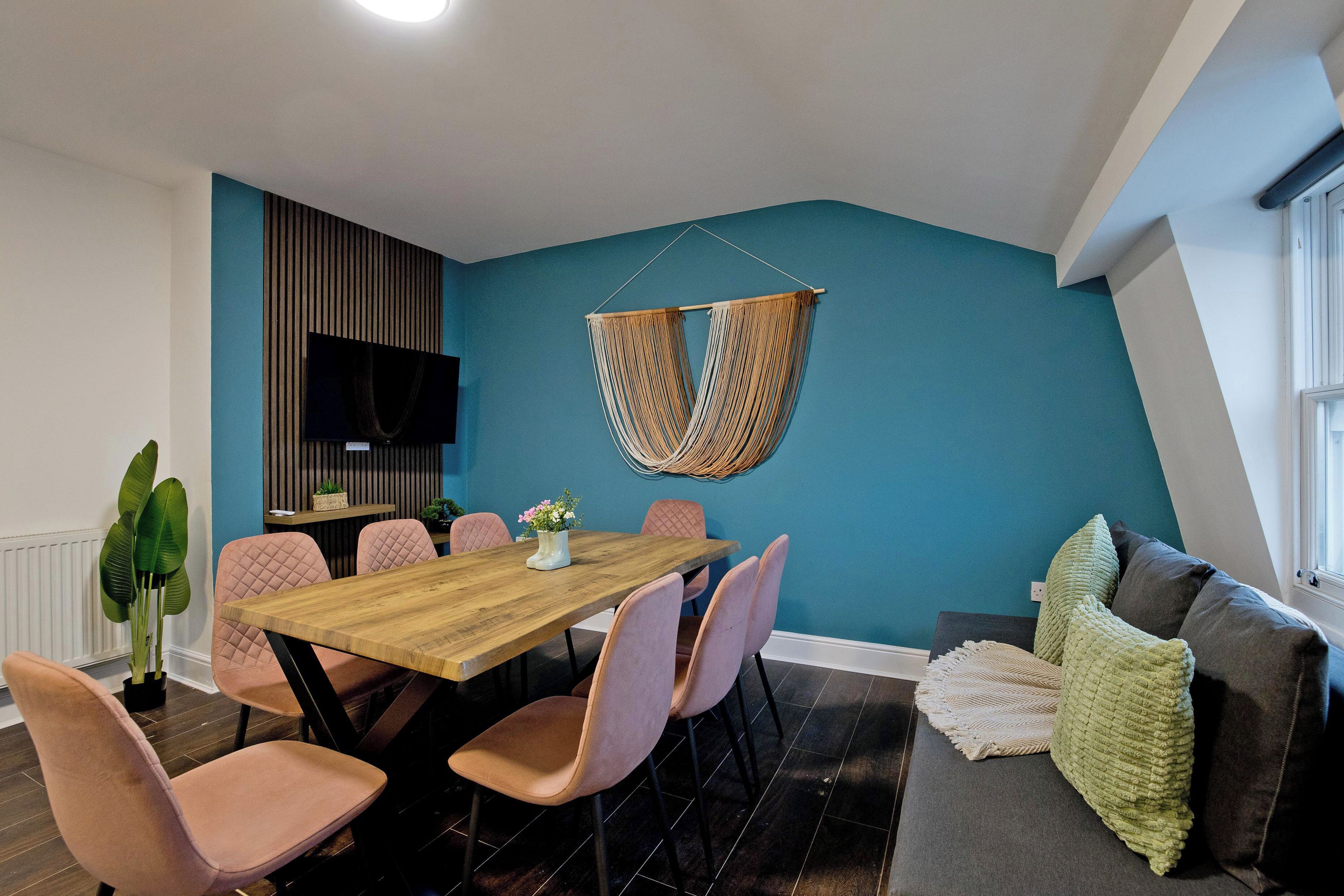 Un comedor con una mesa de madera y sillas rosadas, un sillón colgante y un sofá gris con una manta verde.