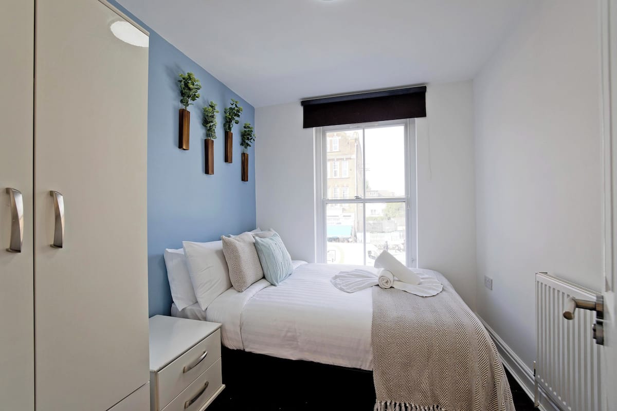 Une chambre à coucher comprenant un lit, des tables de chevet, une armoire et une fenêtre donnant sur des immeubles.