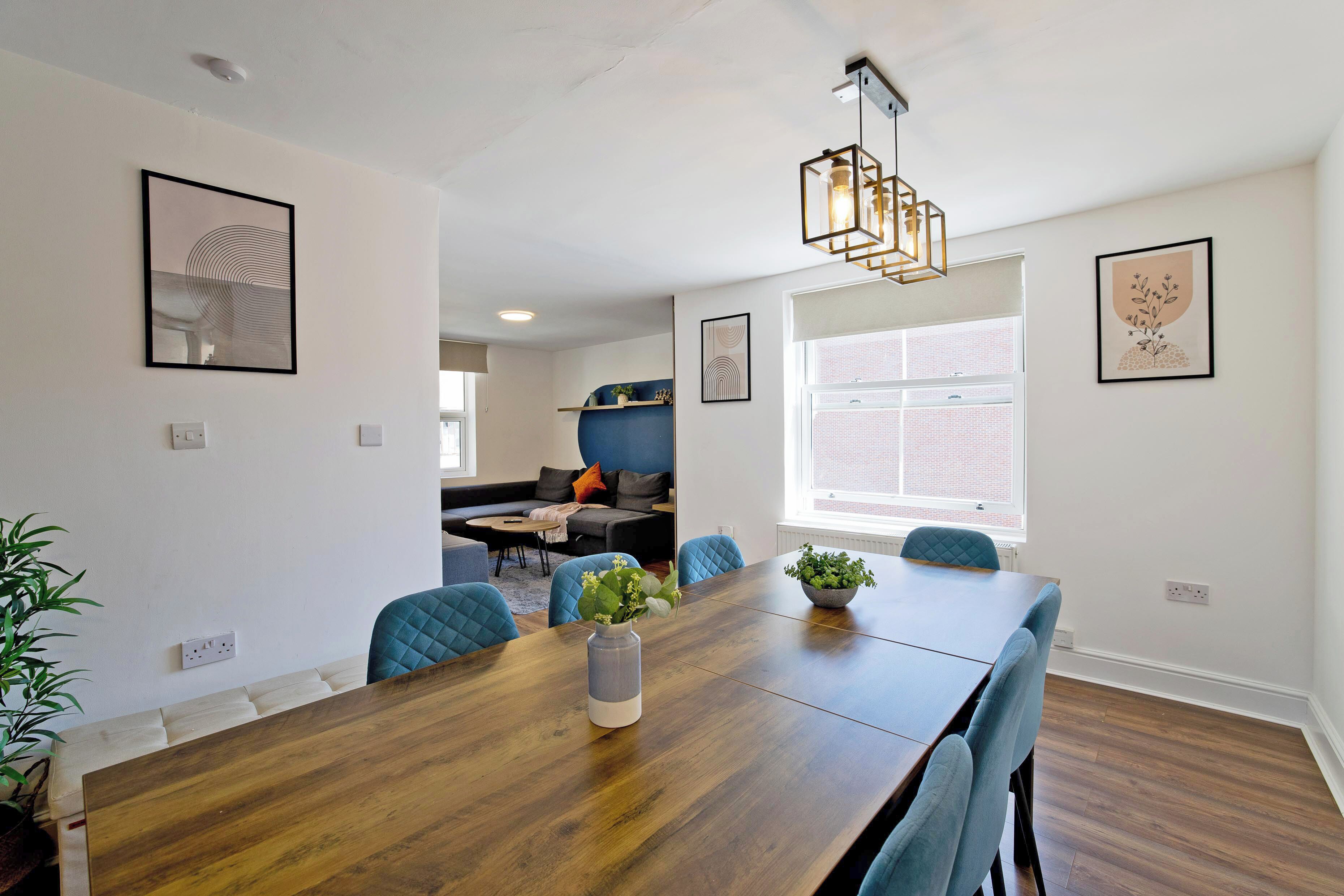 Un coin repas avec une table en bois, des chaises bleues et un vase rempli de fleurs. Les murs sont ornés de tableaux encadrés et un luminaire est suspendu au plafond.