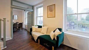 Un salon avec un canapé vert foncé, des coussins blancs, un plancher en bois, un coin repas avec des chaises et une grande fenêtre donnant sur des immeubles et des arbres.