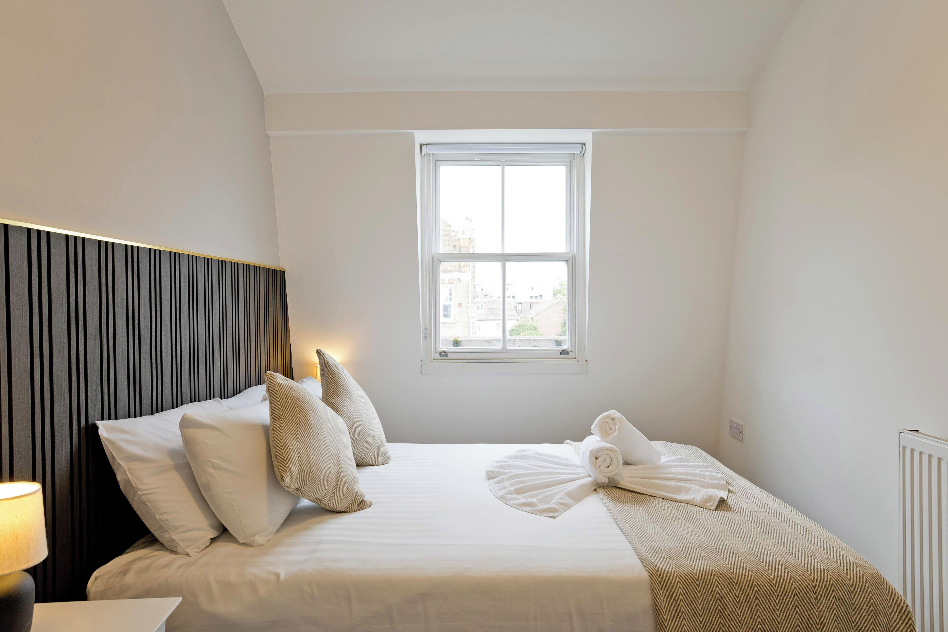 A neatly made bed with white linens and a striped blanket, a bedside lamp, and a window with a view of buildings outside.