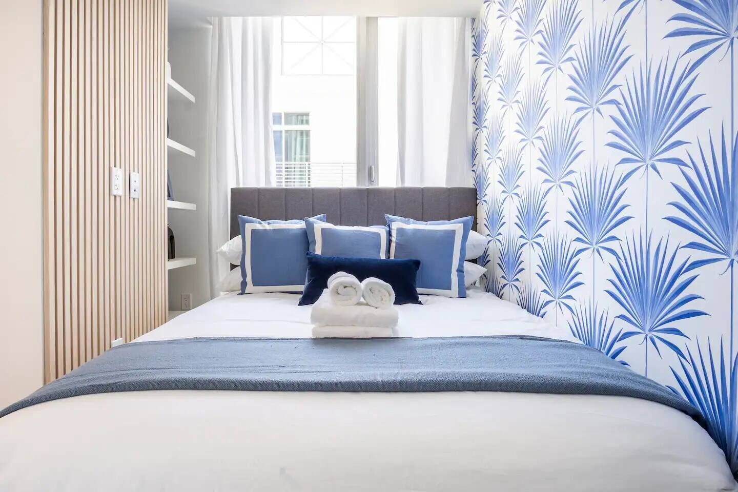A hotel room with a bed, blue and white bedding, a grey headboard, and a window with curtains.