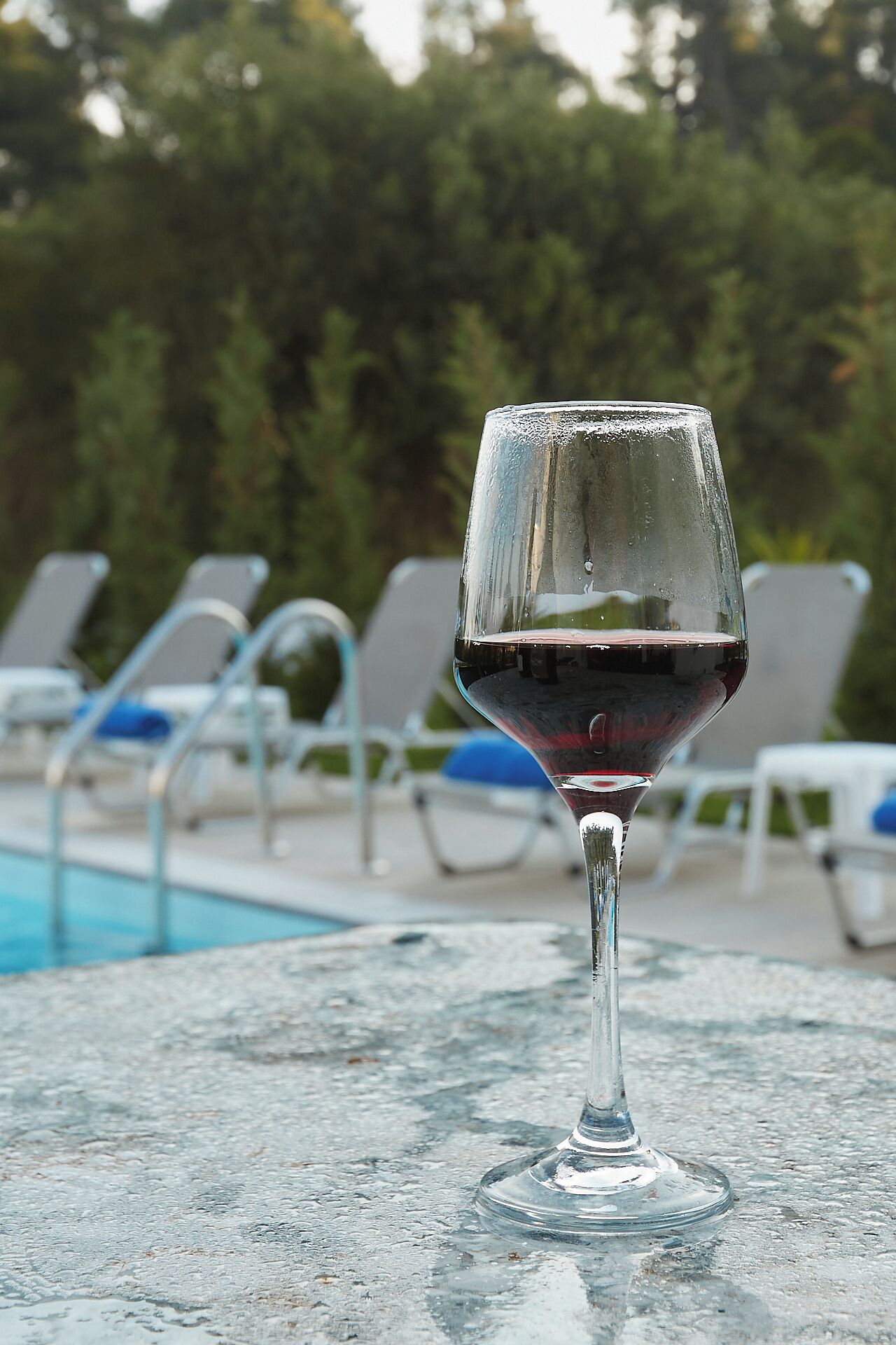A glass of red wine on a table by a poolside with lounge chairs in the background.