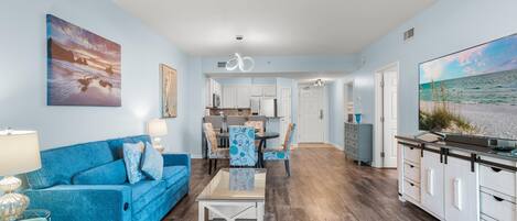 A modern living room with a blue sofa, a dining area, and a kitchen with white cabinets.