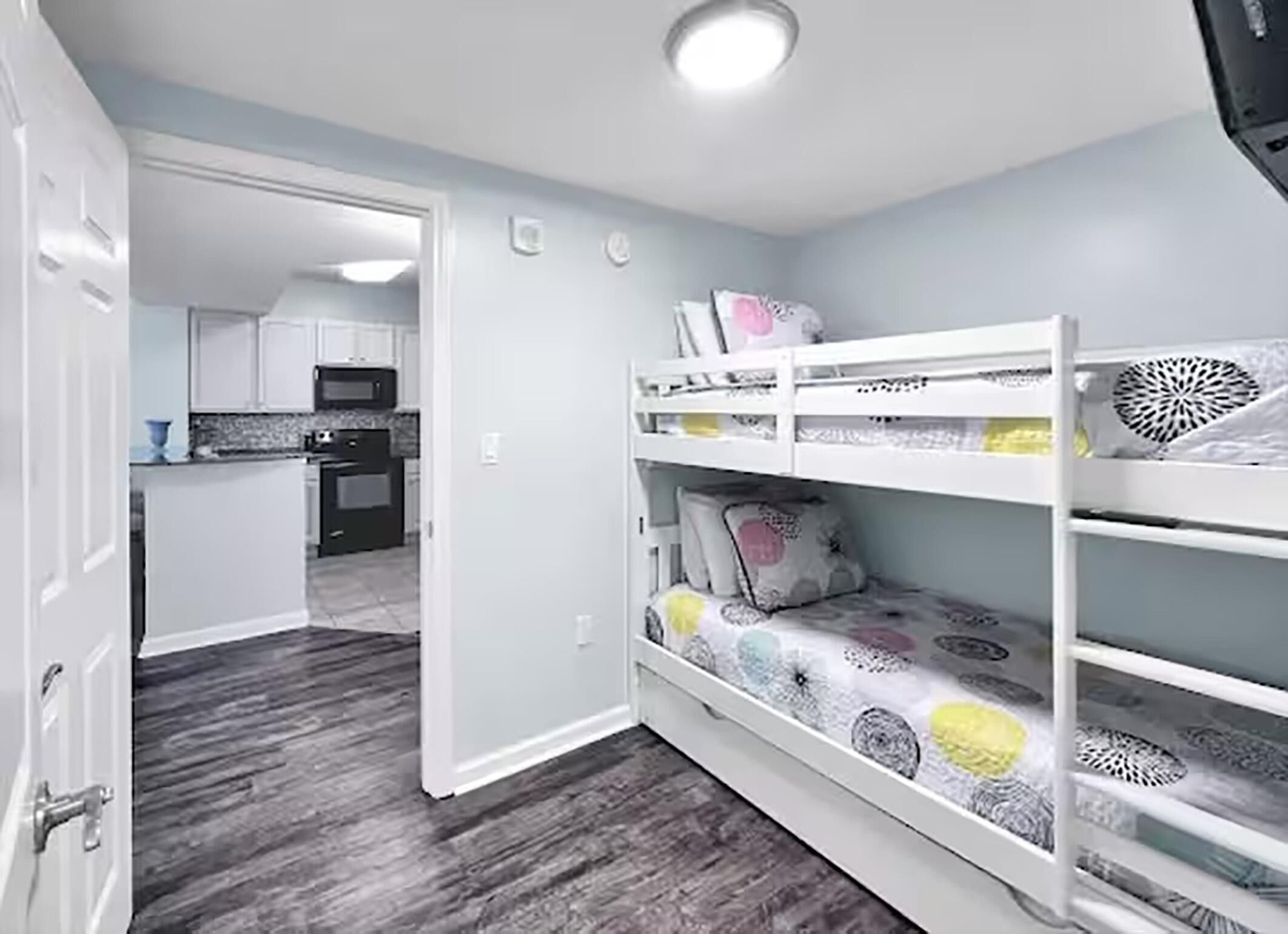 A room with a bunk bed, a kitchen in the background, and a door leading to another room.