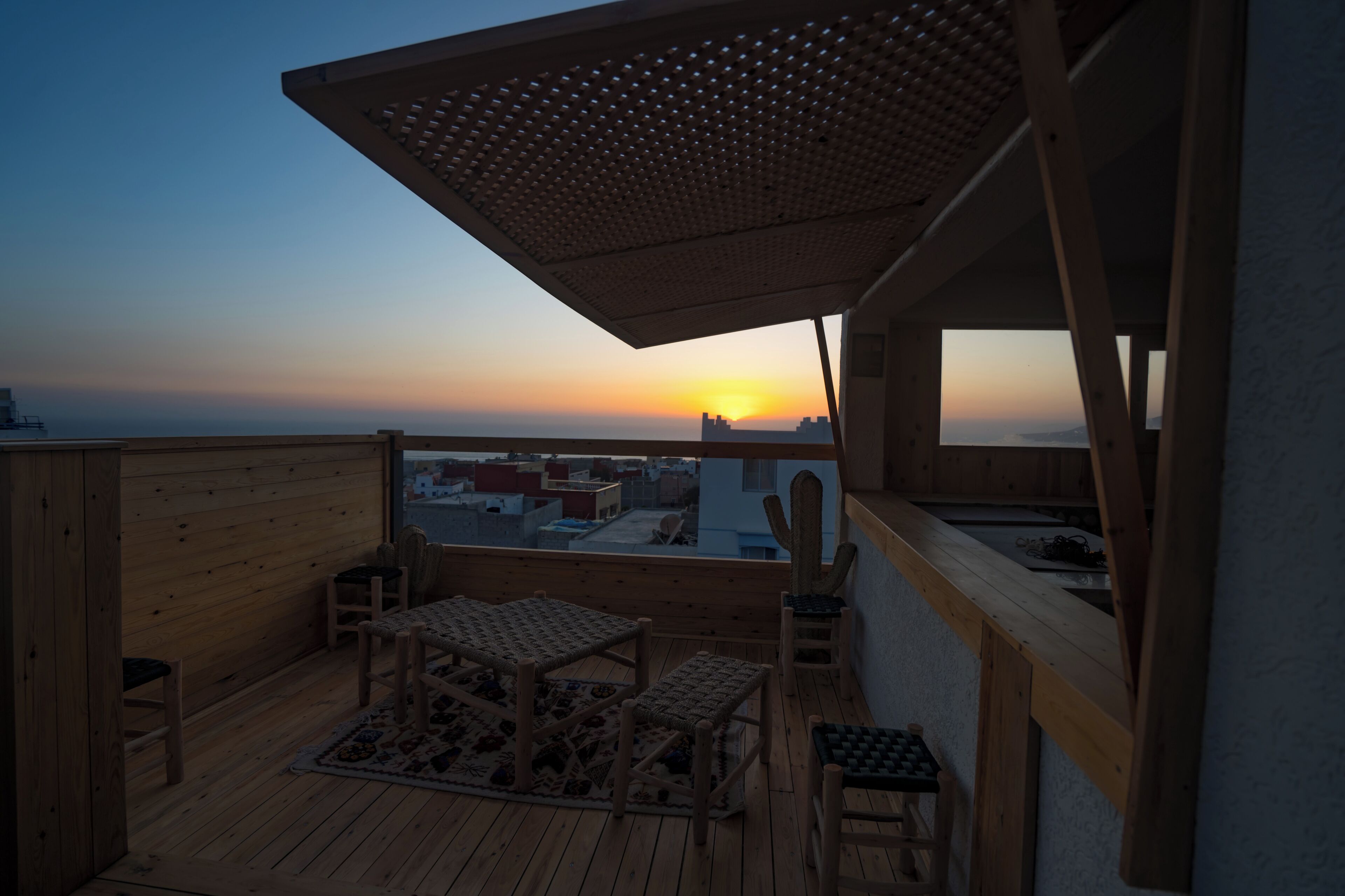 A wooden deck with a table and chairs, a cactus, and a view of a sunset over a cityscape.