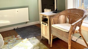 A room with a flat-screen TV mounted on the wall, a wicker chair, a desk with drawers, a radiator, and a window with a view of the outside.