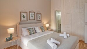 A neatly made bed with white linens and rolled towels, a bedside table with a lamp, and a wall with framed botanical prints.
