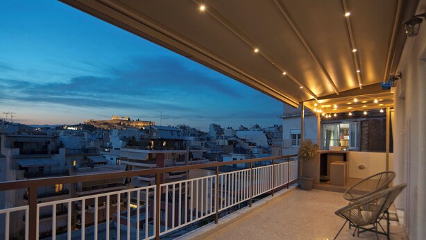 A balcony with a view of a cityscape at dusk, featuring a railing and a seating area with chairs.