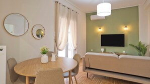 A modern living room with a round dining table, two chairs, a sofa, a TV mounted on the wall, and an air conditioning unit.
