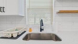 A kitchen with white cabinets, a stainless steel sink, a black faucet, and a window with blinds. - Charming Retreat w/ Bikes (Green Cove Springs)