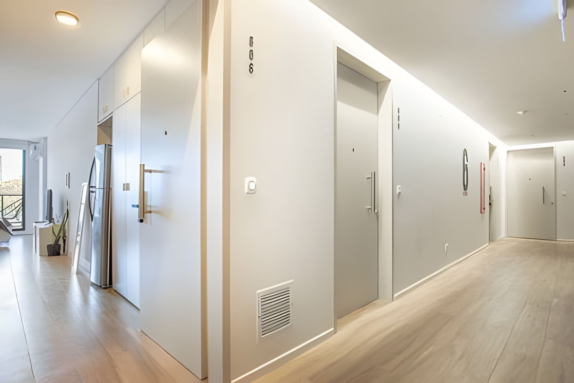 Un passadís modern amb un disseny minimalista, amb combinació de parets blanques i terra de fusta. Hi ha diverses portes i un frigorífic visibles, cosa que suggereix un entorn residencial o d'hotel.