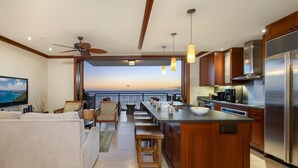 A modern kitchen with a large island, wooden cabinets, and a view of the ocean at sunset.