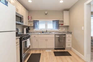 A kitchen with white cabinets, a microwave, stove, and dishwasher. There is a window with patterned curtains, a sink, and a countertop with a soap dispenser.