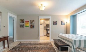 A room with a bed, a bench, a rug, a door, and a kitchen visible through an open door.