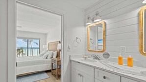 Un baño moderno con un espejo grande, muebles blancos y vista al océano a través de una puerta.