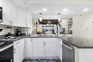 A modern kitchen with white cabinets, granite countertops, a built-in oven, and a dishwasher. - Loggerhead Cay 203 | Reimagined 2BR/2BA Gulf-View Condo with Luxe Amenities (Sanibel)