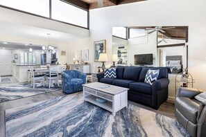A spacious living room with a dark blue sofa, a white coffee table, and a blue armchair. A large window provides natural light, and the room features a marble-patterned rug. - Loggerhead Cay 203 | Reimagined 2BR/2BA Gulf-View Condo with Luxe Amenities (Sanibel)