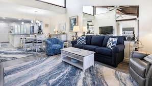 Un salon spacieux avec un canapé bleu foncé, une table basse blanche et un fauteuil bleu. Une grande fenêtre laisse entrer la lumière naturelle, et la pièce est agrémentée d’un tapis à motifs de marbre.