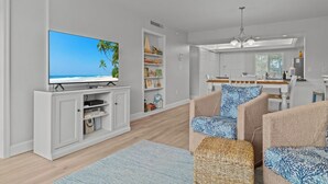 A modern living room with a flat-screen TV, a bookshelf, two patterned armchairs, a wicker ottoman, and a kitchen area with a dining table and chairs.