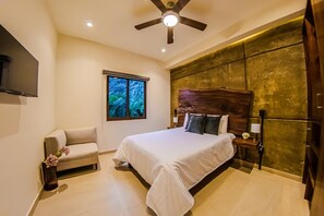 Une chambre à coucher comprenant un lit, une table de chevet, une lampe, un ventilateur de plafond, une fenêtre donnant sur des arbres et un mur avec un motif texturé.
