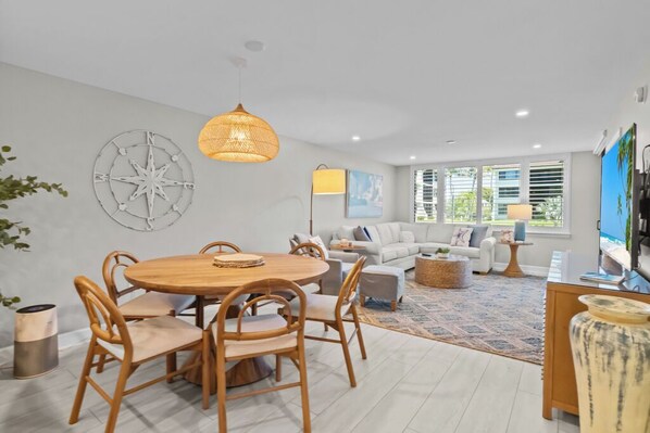 A modern living room with a round wooden dining table, light-colored sofas, a large window with blinds, and a decorative wall piece.