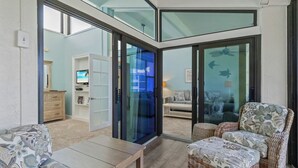A modern living room with a glass door leading to a balcony, wicker furniture, and a television in the background.
