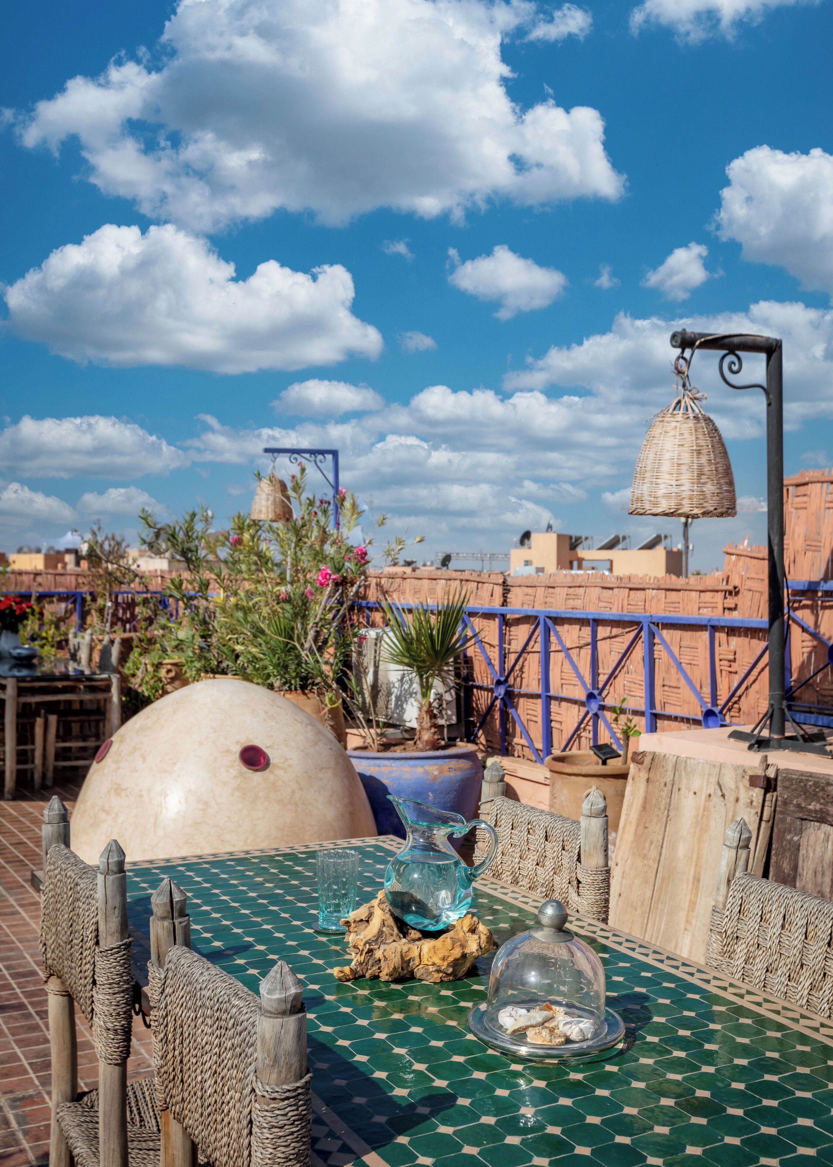A rooftop terrace with a green patterned table, wicker furniture, a large decorative bowl, and a hanging basket.
